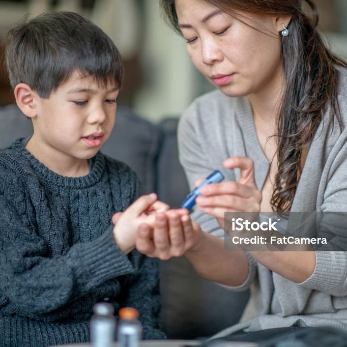 A young Asian boy sits on a sofa beside his mother as she tests his blood sugar level with a lancing device. She is holding his finger out with one hand and has the lancing device in the other. They are both dressed casually.