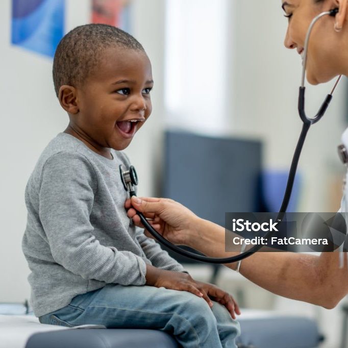 A young boy of African decent, sits up on an exam table as a female Paediatrician preforms a check-up on him.  The boy is dressed casually and smiling as the doctor listens to his heart.