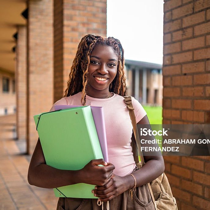 Portrait of a cheerful black female student holding folders and a backpack in a university hallway