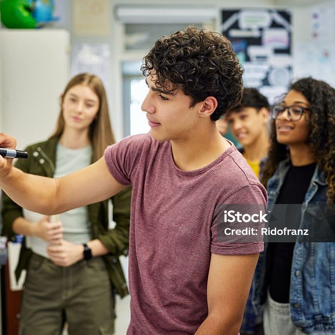 Male student writing on whiteboard while explaining a concept to his classmates at secondary school. Focused teens delivers a group presentation while drawing on board as classmates observe attentively. Teen guy helping classmates understand solution to geometry or arithmetic problem in classroom.
