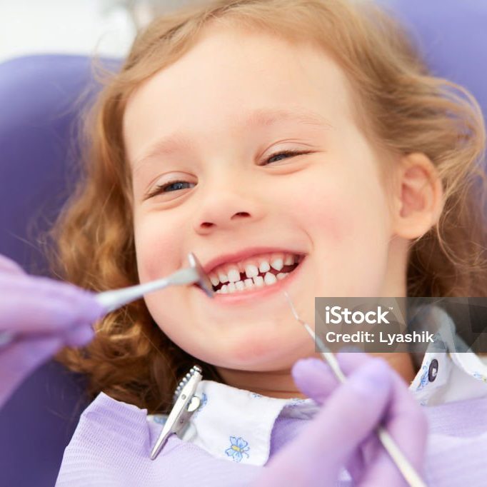 Hands of unrecognizable pediatric dentist making examination procedure for smiling cute little girl sitting on chair in hospital. Dentist office. Little girl sitting in the dentists office